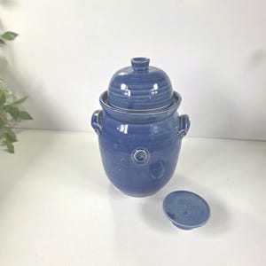 May include: A medium blue ceramic jar with a matching domed lid and two side handles. The jar features a small circular embossed detail on the front. A separate, smaller circular blue ceramic lid or coaster is placed on the surface next to the jar.