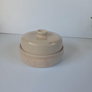 May include: A beige ceramic butter dish with a lid. The dish is round with a slightly flared base and a matching lid with a small knob on top. The glaze has subtle colour variations, giving it a handmade appearance. The butter dish is designed to hold butter.