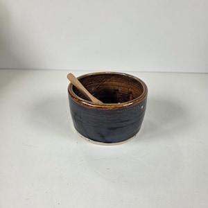 May include: A small, round, dark brown ceramic bowl with a glossy finish and a lighter brown interior rim. A small, light-coloured wooden spoon rests inside the bowl, angled against the inner wall. The base of the bowl is a contrasting light beige.