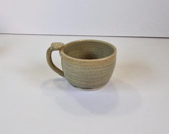 May include: A green ceramic mug with a unique handle. The mug has a speckled glaze and a slightly textured surface.