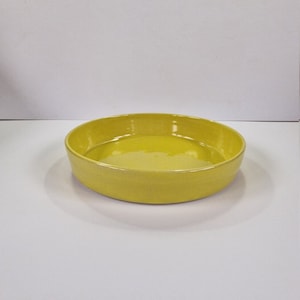 May include: A bright yellow ceramic bowl with a rounded shape. The bowl is empty and sitting on a white surface.