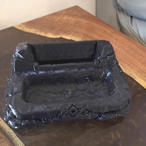 May include: A black, rectangular ashtray with a textured surface and a carved design. The ashtray has a recessed area for holding items and a decorative pattern on the front.