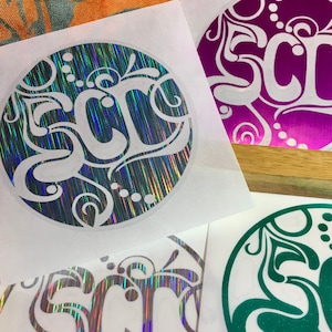 May include: Four round stickers with a white background and a holographic, iridescent, or glitter finish. Each sticker features a white, stylized design of the word "SCR" with a decorative flourish. The stickers are in different colors: blue, purple, green, and silver.