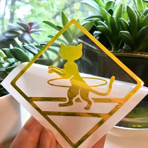 May include: A gold-colored vinyl decal of a cat holding a hula hoop. The decal is on a white background and is shaped like a diamond.