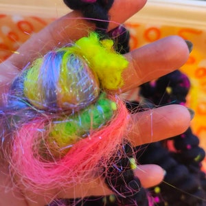 May include: A hand holding a colorful yarn ball with pink, green, yellow, and blue strands. The yarn is wrapped around a black yarn ball.