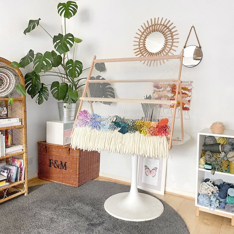 May include: A wooden weaving loom with a rainbow-colored woven tapestry. The tapestry features a variety of colors, including blue, green, yellow, and orange, with a long cream-colored fringe. The loom is on a white pedestal base.