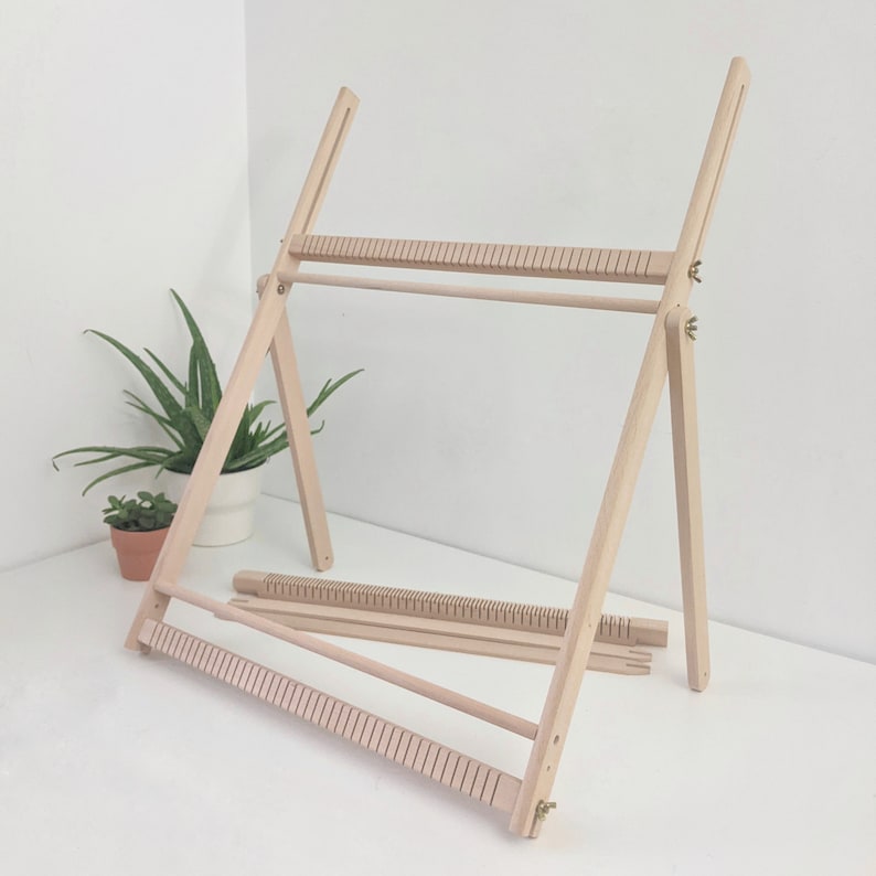 May include: A wooden weaving loom with a natural finish. The loom is adjustable and features a frame with multiple horizontal bars and vertical slots. Additional wooden pieces are included. A small potted plant is in the background.