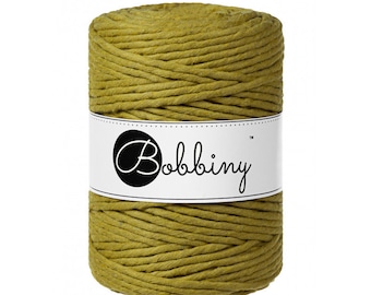 Könnte beinhalten: Eine Spule mit senfgelbem Baumwollgarn, beschriftet mit "Bobbiny" auf einem weißen Etikett.