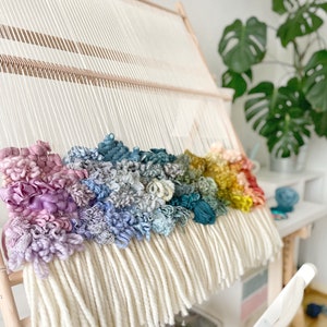 May include: A colorful, handmade woven wall hanging in progress. The piece features a rainbow of textured yarn in shades of pink, blue, green, yellow, and red, set against a cream-colored background. The weaving is mounted on a wooden frame.