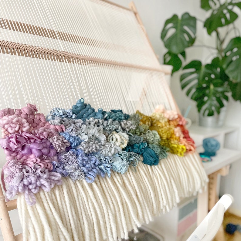 May include: A colorful, handmade woven wall hanging in progress. The piece features a rainbow of textured yarn in shades of pink, blue, green, yellow, and red, set against a cream-colored background. The weaving is mounted on a wooden frame.