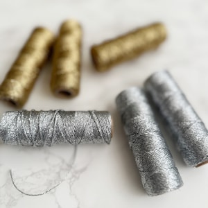 May include: Five spools of metallic thread, two gold and three silver, on a white marble surface.