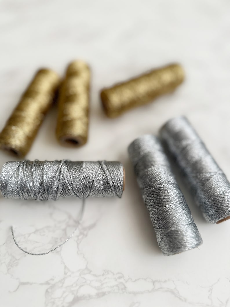 May include: Five spools of metallic thread, two gold and three silver, on a white marble surface.
