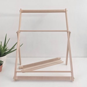 May include: A wooden weaving loom with a natural finish. The loom has a rectangular frame with horizontal bars and vertical supports. Two additional wooden pieces are included. The loom is designed for textile art and craft projects.