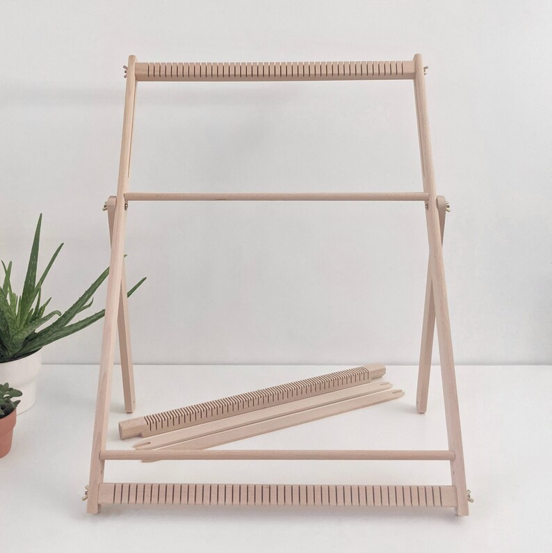 May include: A wooden weaving loom with a natural finish. The loom has a rectangular frame with horizontal bars and vertical supports. Two additional wooden pieces are included. The loom is designed for textile art and craft projects.