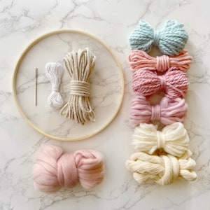 May include: A set of yarn and rope for macrame wall hanging craft projects. The set includes a wooden hoop, a needle, white string, beige rope, and five different colours of yarn in shades of pink, blue, and cream.