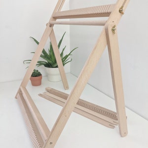 May include: A wooden weaving loom with a natural finish, set against a white background. The loom is in an A-frame design, with a horizontal bar and several weaving tools. A potted plant is visible in the background.