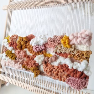 May include: A colorful woven wall hanging in shades of pink, peach, mustard yellow, and white. The textured piece is mounted on a wooden loom, showcasing various weaving techniques and yarn textures. The artwork is a unique home decor item.