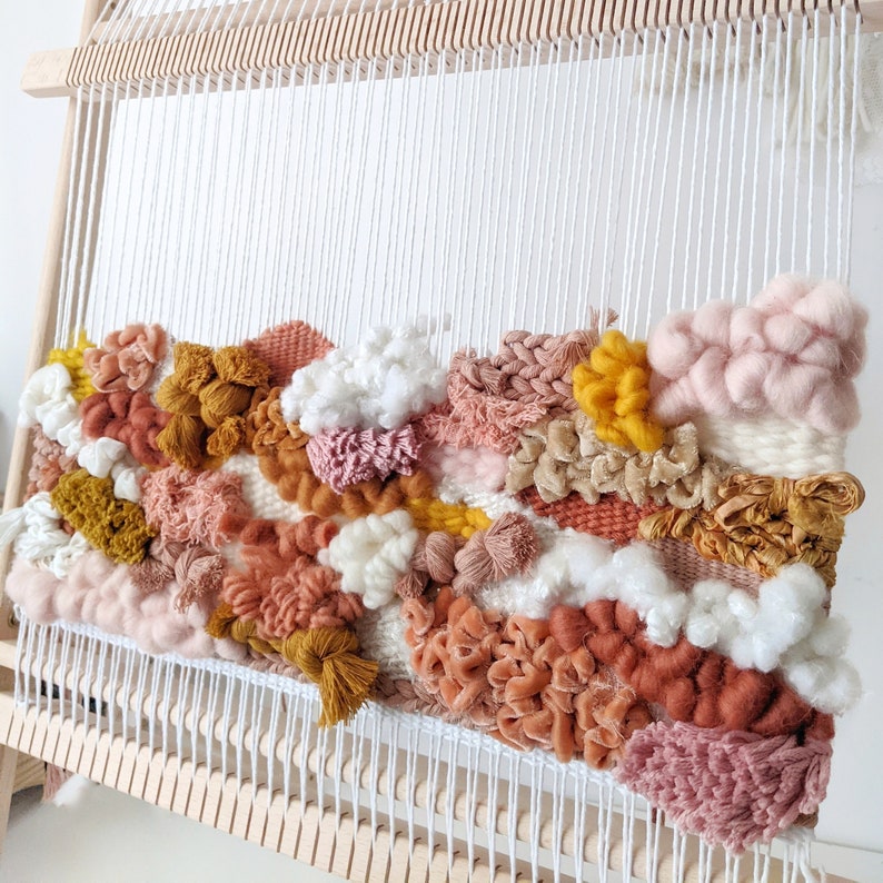 May include: A colorful woven wall hanging in shades of pink, peach, mustard yellow, and white. The textured piece is mounted on a wooden loom, showcasing various weaving techniques and yarn textures. The artwork is a unique home decor item.