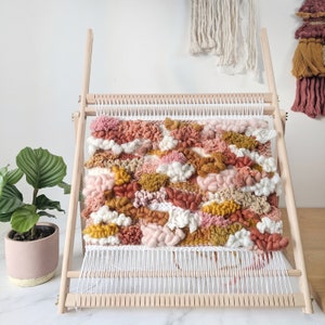 May include: A woven wall hanging in a wooden frame. The textile art piece features a textured design with various shades of pink, white, brown, and yellow. The weaving is displayed on a wooden loom, with a potted plant in the background.