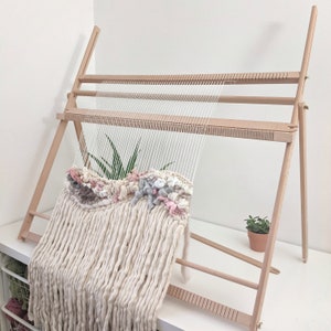 May include: A wooden weaving loom with a partially completed textile project. The textile features cream-colored, chunky yarn with accents of pink, gray, and white. The loom is set up on a white surface, with a small potted plant visible.