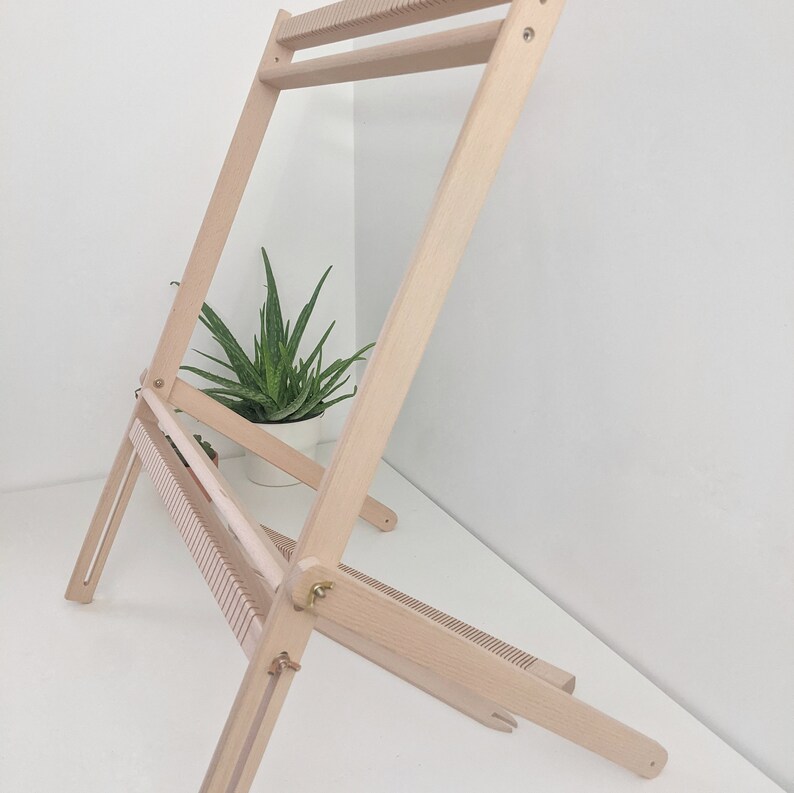 May include: A light-colored wooden weaving loom, angled and partially folded. The loom has multiple horizontal slots and a top bar. A small potted plant is visible in the background.
