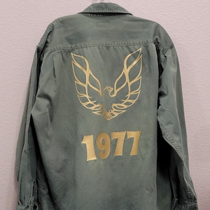 May include: An olive green button-down shirt with a gold graphic of a bird and the number 1977 printed on the back. The shirt is hanging on a metal hook against a light pink wall.