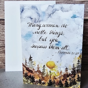 May include: A greeting card with a watercolor painting of a sunset over a forest. The card has the text "Many women do noble things, but you surpass them all. Proverbs 31:29". A white envelope is next to the card.