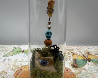 May include: A glass jar with a cork base contains a unique assemblage. A human tooth is suspended from the top, connected to a blue skull bead and wooden beads. At the bottom, an eye sculpture is surrounded by green moss.