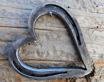Könnte beinhalten: Eine herzförmige Metallskulptur aus einem Hufeisen. Die Skulptur ist in einem rustikalen, dunkelgrauen Farbton gehalten und auf einer Holzoberfläche ausgestellt.