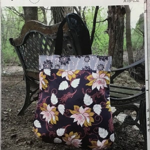 May include: A floral tote bag with black, white, pink, and yellow flowers on a dark purple background. The tote bag is sitting on a wooden bench in a wooded area. The tote bag is 19 inches high, 17 1/2 inches wide, and 3 inches deep. The Soho Tote ATB-152.