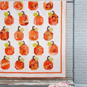 May include: A patchwork quilt featuring a pattern of orange and white pumpkins. The quilt is displayed on a wooden bench with a white brick wall in the background. The text "Patch Work" and "Fresh designs from a Wyoming farm." is displayed below the quilt.