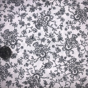 May include: White fabric with a repeating black floral pattern. The design features bouquets of roses, bows, and vines. A quarter coin is visible in the lower left corner for scale. The fabric is likely used for sewing or crafting projects.