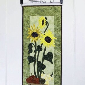 May include: A green quilted wall hanging with a brown wheelbarrow filled with sunflowers, gourds, and apples. The wheelbarrow is on a green background with a white border.