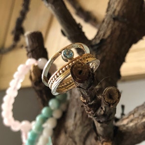 May include: A silver and gold ring with a blue gemstone set in the center. The ring is on a branch with a pink and green beaded bracelet in the background.