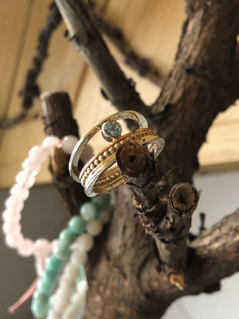 May include: A silver and gold ring with a blue gemstone set in the center. The ring is on a branch with a pink and green beaded bracelet in the background.