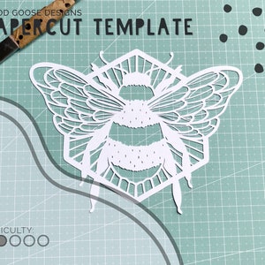 May include: A white papercut template of a bee in a hexagon shape. The bee has detailed wings and a striped body. The template is on a green cutting mat with a difficulty rating of two out of five.