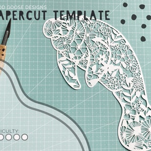 May include: A white papercut template of a manatee with intricate floral designs. The template is on a green cutting mat with a craft knife nearby. The difficulty level is indicated by five circles, all filled in.