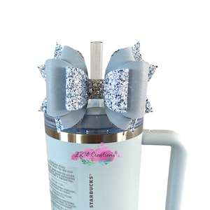 May include: A light blue stainless steel tumbler with a handle and a white lid. The tumbler has a silver band around the top and a white sticker with the text "STANLEY + STARBUCKS" and "ZRM Creations". A light blue and silver glitter bow with a clear straw is on top of the tumbler.