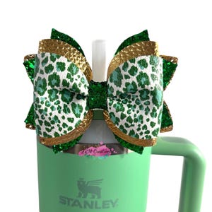 Puede incluir: Un vaso Stanley Quencher H2.0 verde con un asa a juego. El vaso está rematado con un gran lazo con estampado de leopardo verde y blanco, ribete dorado y detalles de purpurina verde. Una pajita blanca está adjunta.