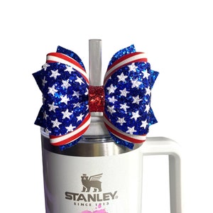 May include: A white stainless steel travel mug with a red, white, and blue glitter bow on top. The mug has the Stanley logo and the text "Since 1913". The mug also has the text "ZRM Creations" in pink.