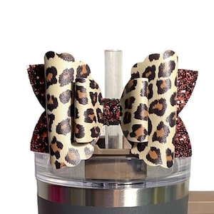 Puede incluir: Una taza de viaje de acero inoxidable gris con un asa marrón. La taza tiene un diseño de estampado de leopardo alrededor de la parte inferior y un lazo de estampado de leopardo con un acento de purpurina roja en la tapa. La taza tiene el logotipo de Stanley y el texto "ZRM Creations" en el lateral.