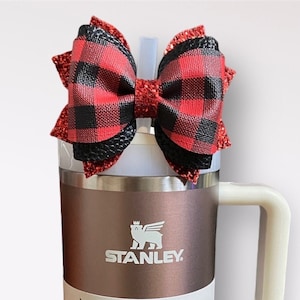 May include: A stainless steel Stanley Quencher H2.0 Tumbler with a black and red plaid bow on top. The tumbler is 40 ounces and has a white handle. The text "STANLEY" is printed on the side of the tumbler. The text "THE FLOWSTATE" is printed above the text "QUENCHER H2.0 TUMBLER". The text "40 OZ | 1.18 L" is printed below the text "QUENCHER H2.0 TUMBLER". The text "All Day Hydration" is printed below the text "40 OZ | 1.18 L". The text "ZRM Creations" is printed on the bottom of the tumbler.