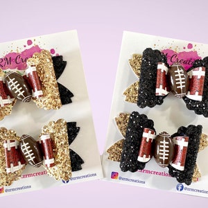 May include: Two pairs of hair bows with a football design. One pair is black and gold glitter, the other is black glitter. Both pairs feature a brown football with white laces.