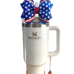 May include: White stainless steel travel mug with a red and blue glitter bow on the lid. The mug has a handle and a silver logo that says "STANLEY SINCE 1913". A small red, white, and blue charm hangs from the handle.