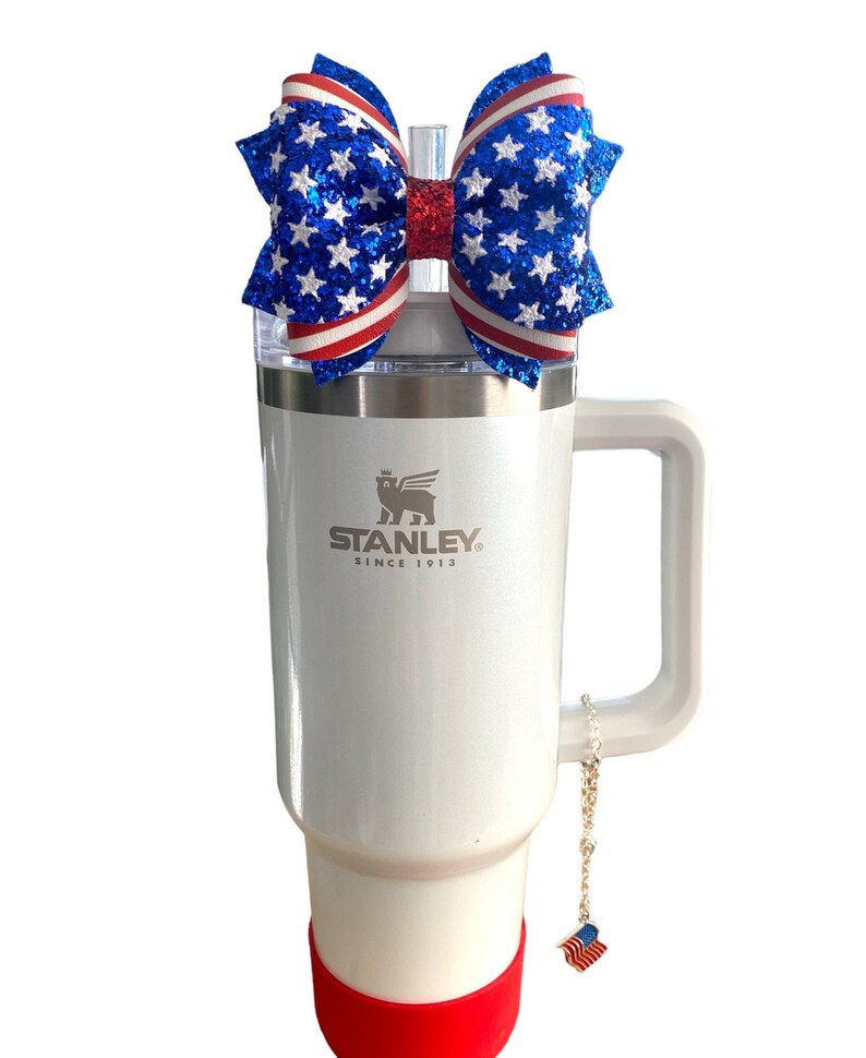 May include: White stainless steel travel mug with a red and blue glitter bow on the lid. The mug has a handle and a silver logo that says "STANLEY SINCE 1913". A small red, white, and blue charm hangs from the handle.