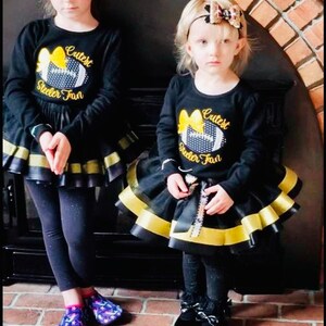 May include: Two girls wearing black long-sleeved shirts with a yellow and black football design and the text "Cutest Steeler Fan". They are wearing black and gold layered skirts and black leggings. One girl is wearing black shoes and the other is wearing purple and pink socks.