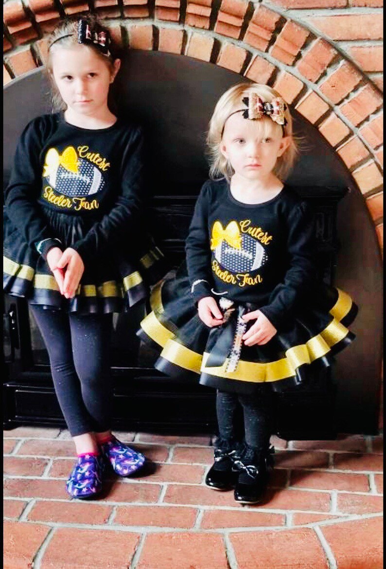 May include: Two girls wearing black long-sleeved shirts with a yellow and black football design and the text "Cutest Steeler Fan". They are wearing black and gold layered skirts and black leggings. One girl is wearing black shoes and the other is wearing purple and pink socks.