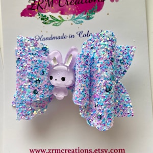 K&ouml;nnte beinhalten: Eine lila Glitzer-Haarbogen mit einem Lavendel-Hasen-Anh&auml;nger. Die Schleife ist aus schimmerndem Glitzer gefertigt und hat eine wei&szlig;e Basis.