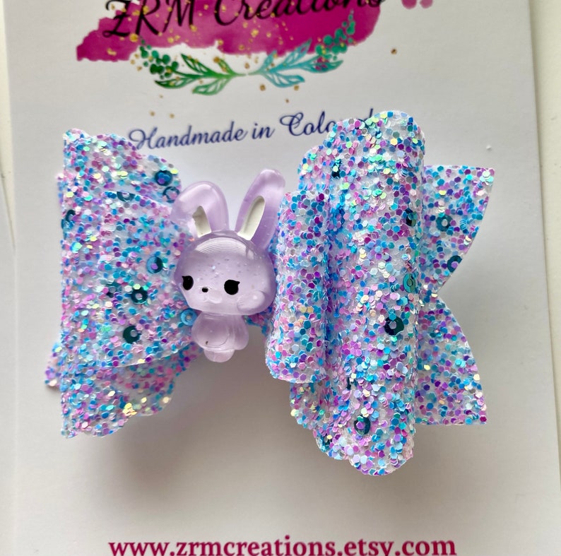 K&ouml;nnte beinhalten: Eine lila Glitzer-Haarbogen mit einem Lavendel-Hasen-Anh&auml;nger. Die Schleife ist aus schimmerndem Glitzer gefertigt und hat eine wei&szlig;e Basis.