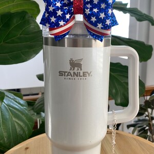 May include: White Stanley travel mug with a red, white, and blue glitter bow on top. The mug has a handle and a red silicone base. The mug has the Stanley logo and the text "Since 1913". A silver chain with a small American flag charm hangs from the handle.